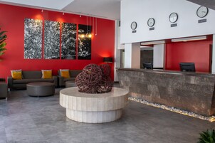 Een moderne receptiezone met een rode muur, een groot abstract schilderij, een receptiebalie en zitmogelijkheden met een opvallende, bolvormige sculptuur.