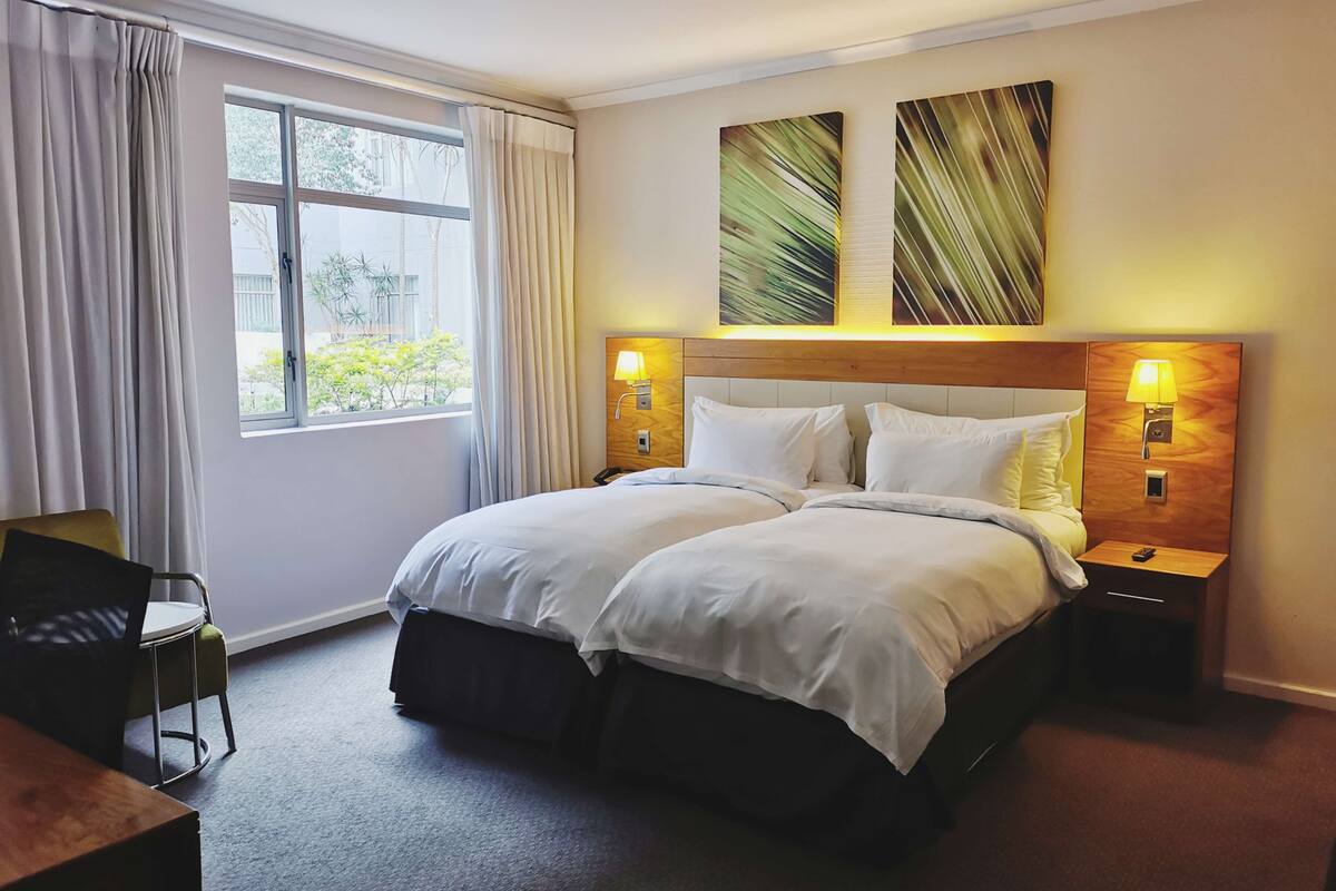 Een hotelkamer met een tweepersoonsbed, twee bedlampen, een raam met gordijnen, een kleine tafel met een stoel, en twee decoratieve schilderijen.