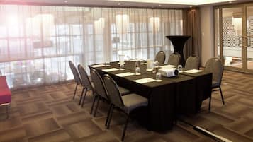 Un salón de conferencias con una larga mesa, sillas, un proyector y un ventanal con cortinas translúcidas.