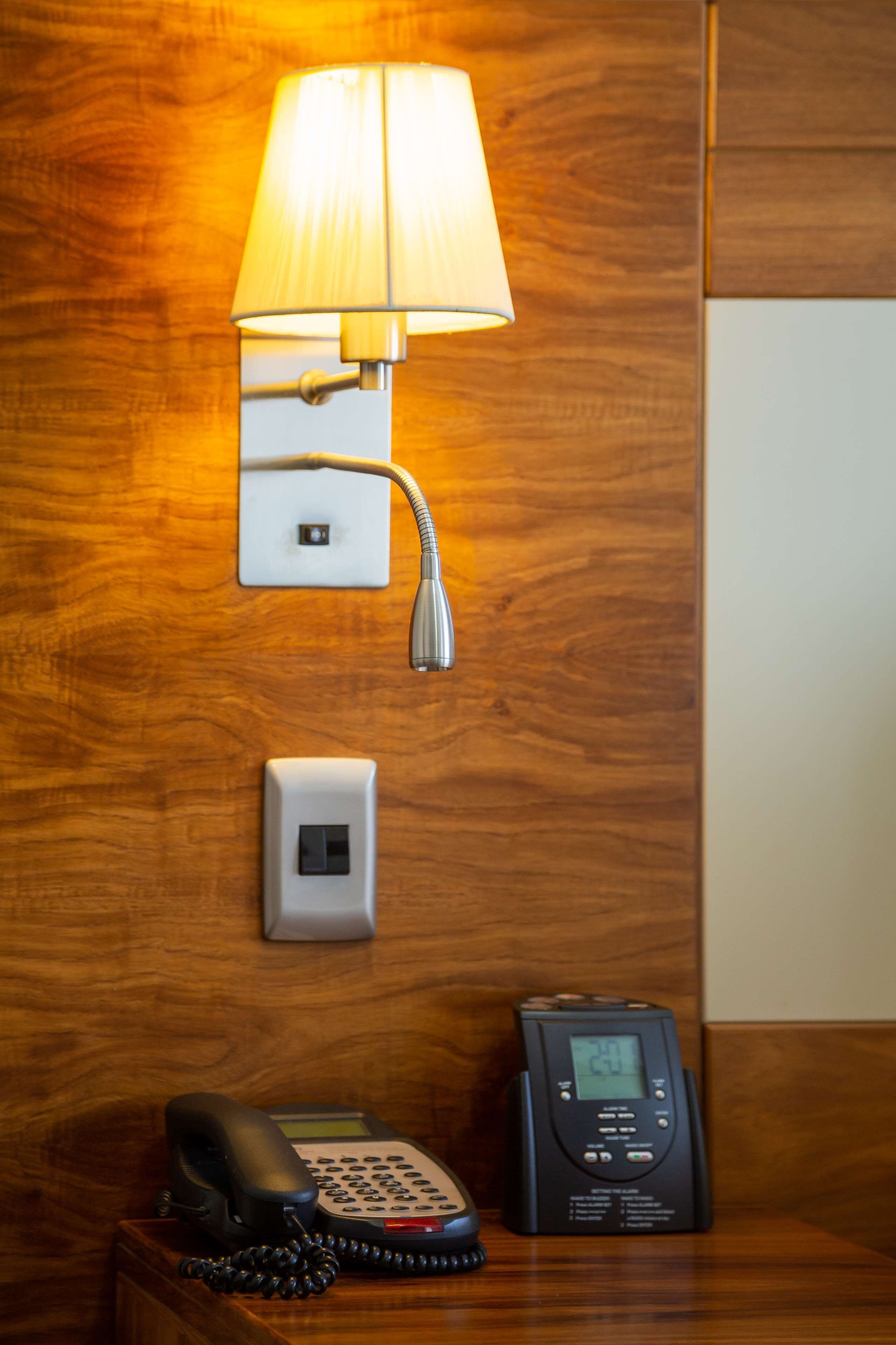 A hotel room with a wooden wall, a wall-mounted telephone, a bedside table with a lamp, and a digital clock.