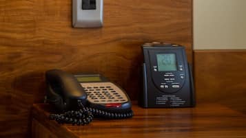 A hotel room with a wooden wall, a wall-mounted telephone, a bedside table with a lamp, and a digital clock.