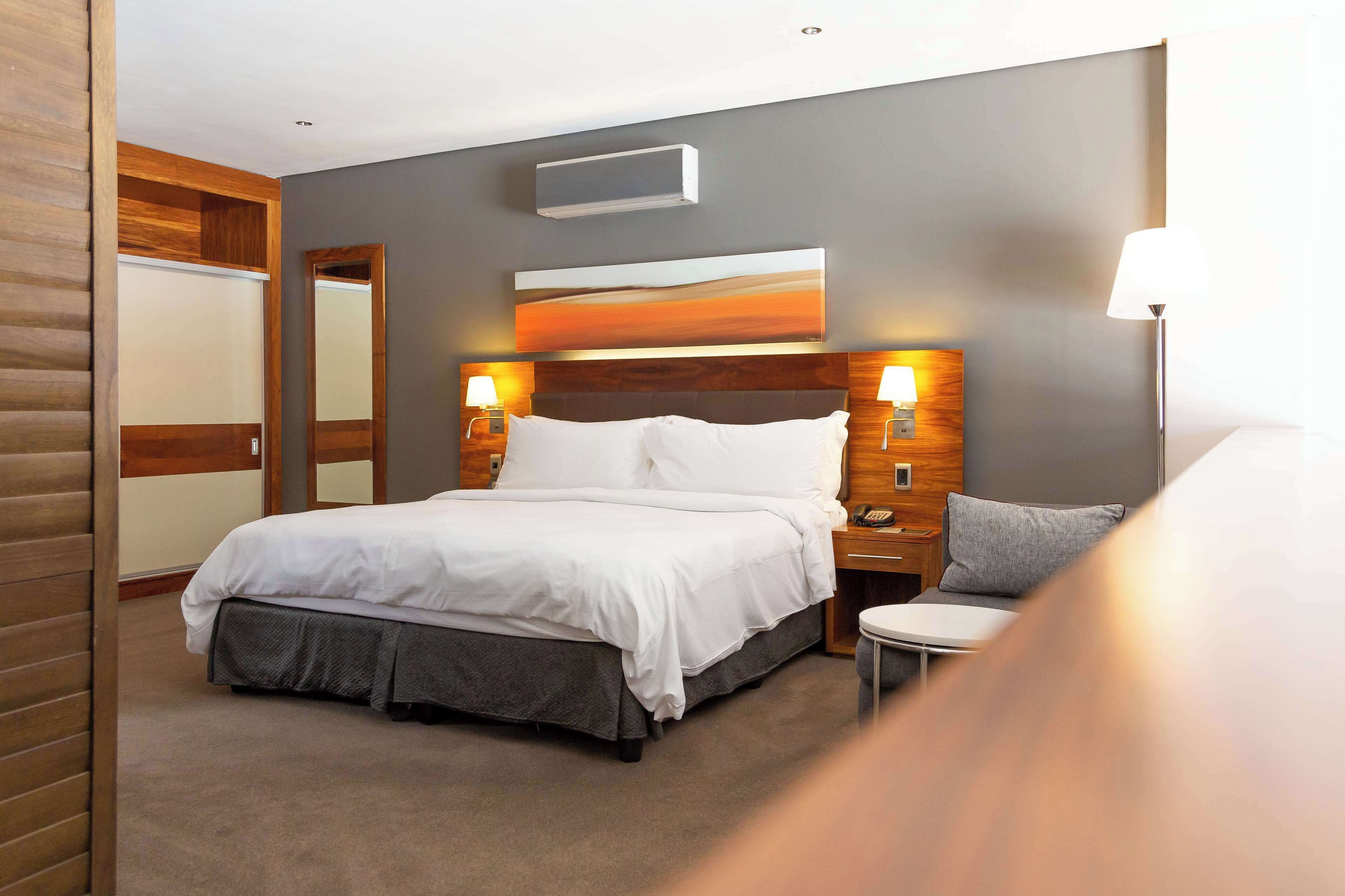 A modern hotel room with a large bed, two bedside tables with lamps, a wall-mounted air conditioner, and a large mirror.