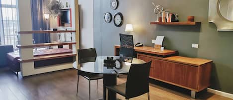 A modern hotel room with a dining area featuring a round glass table and black chairs, a wooden desk with a lamp, and a bookshelf with decorative items.