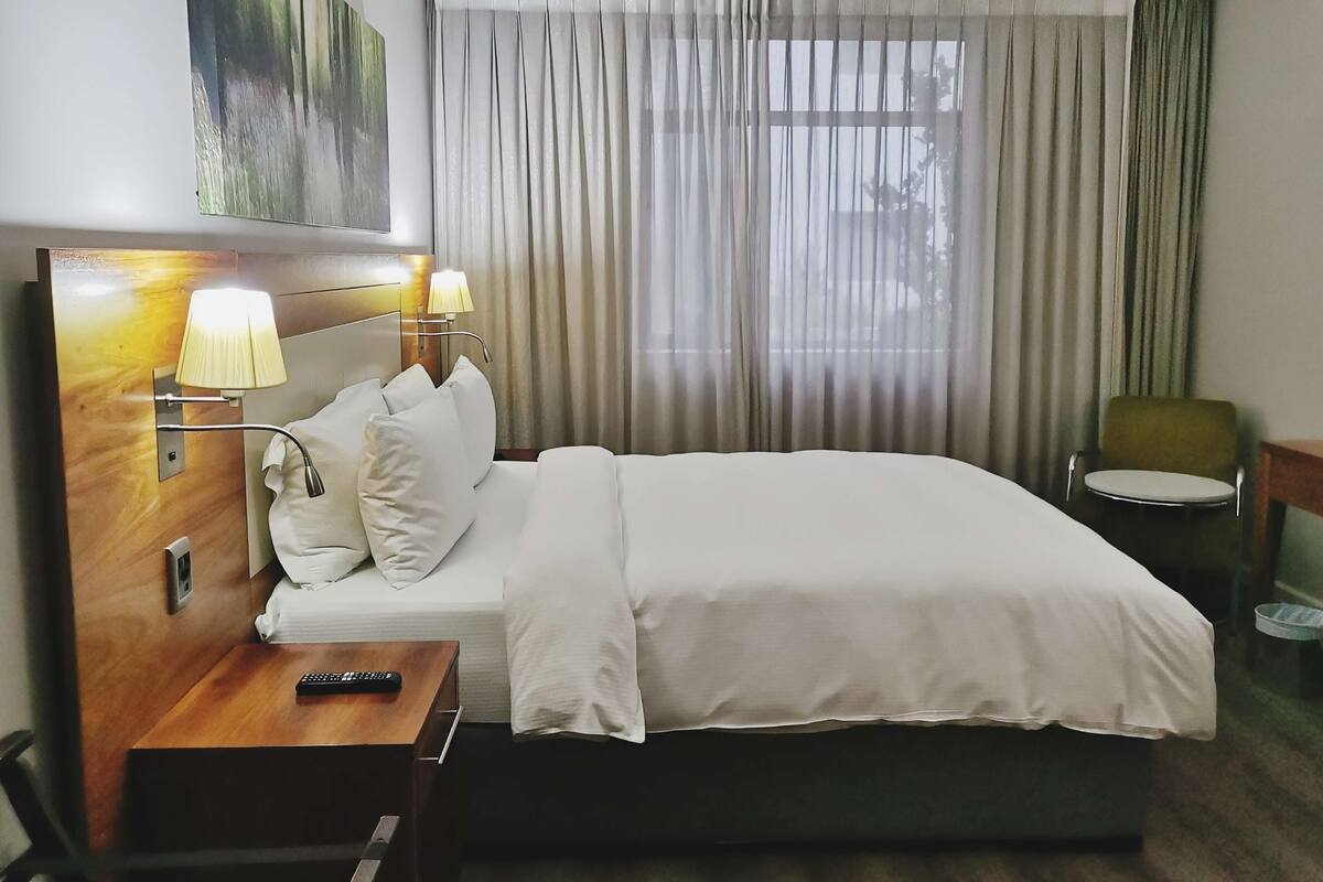 Hotelkamer met een bed, bedlampen, nachtkastje, afstandsbediening, stoel en raam met gordijnen.