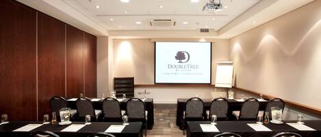 A conference room with a projector screen displaying "DoubleTree by Hilton Cape Town - Upper Eastside" logo, multiple tables with chairs, and a whiteboard.
