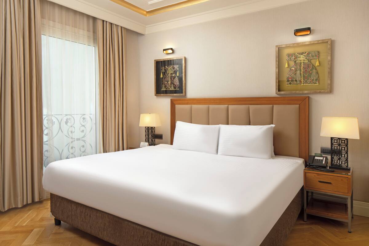 A hotel room with a large bed, two bedside tables with lamps, a framed artwork on the wall, and a window with curtains.
