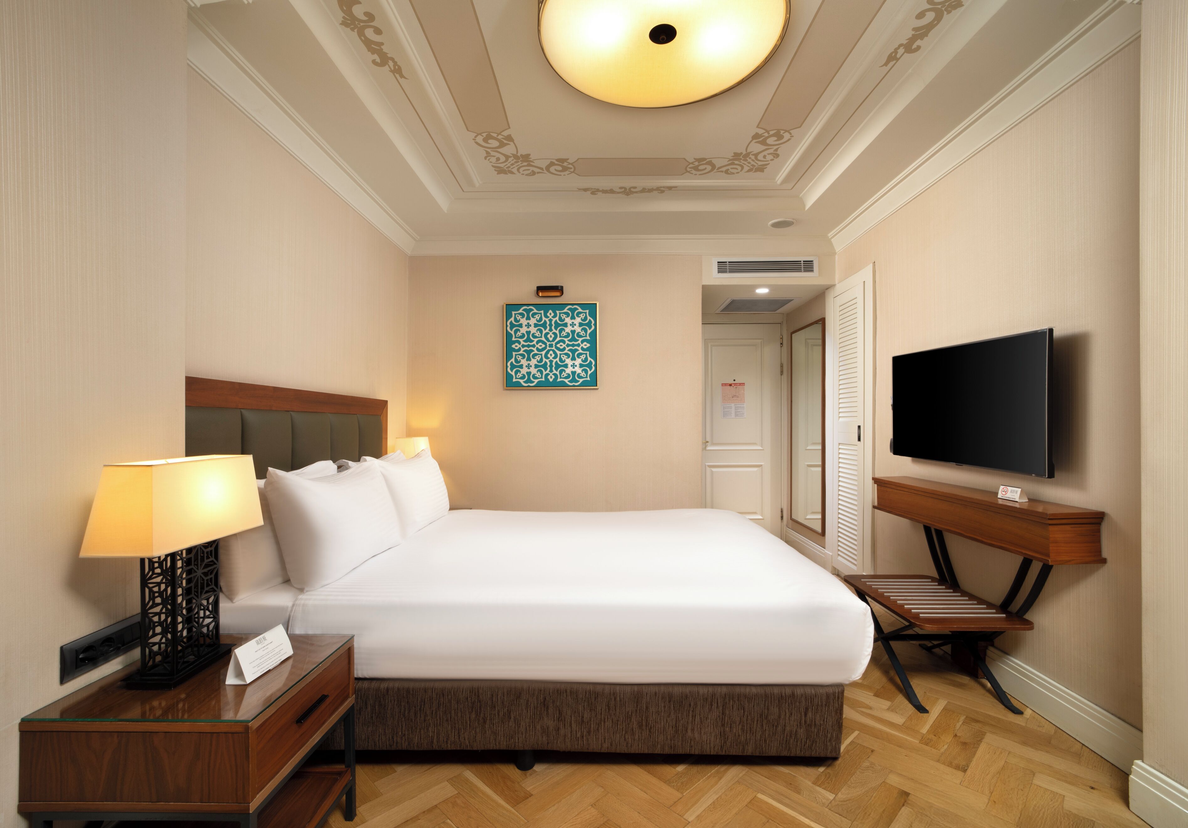 A hotel room with a large bed, a bedside table with a lamp, a TV mounted on the wall, and a decorative ceiling.