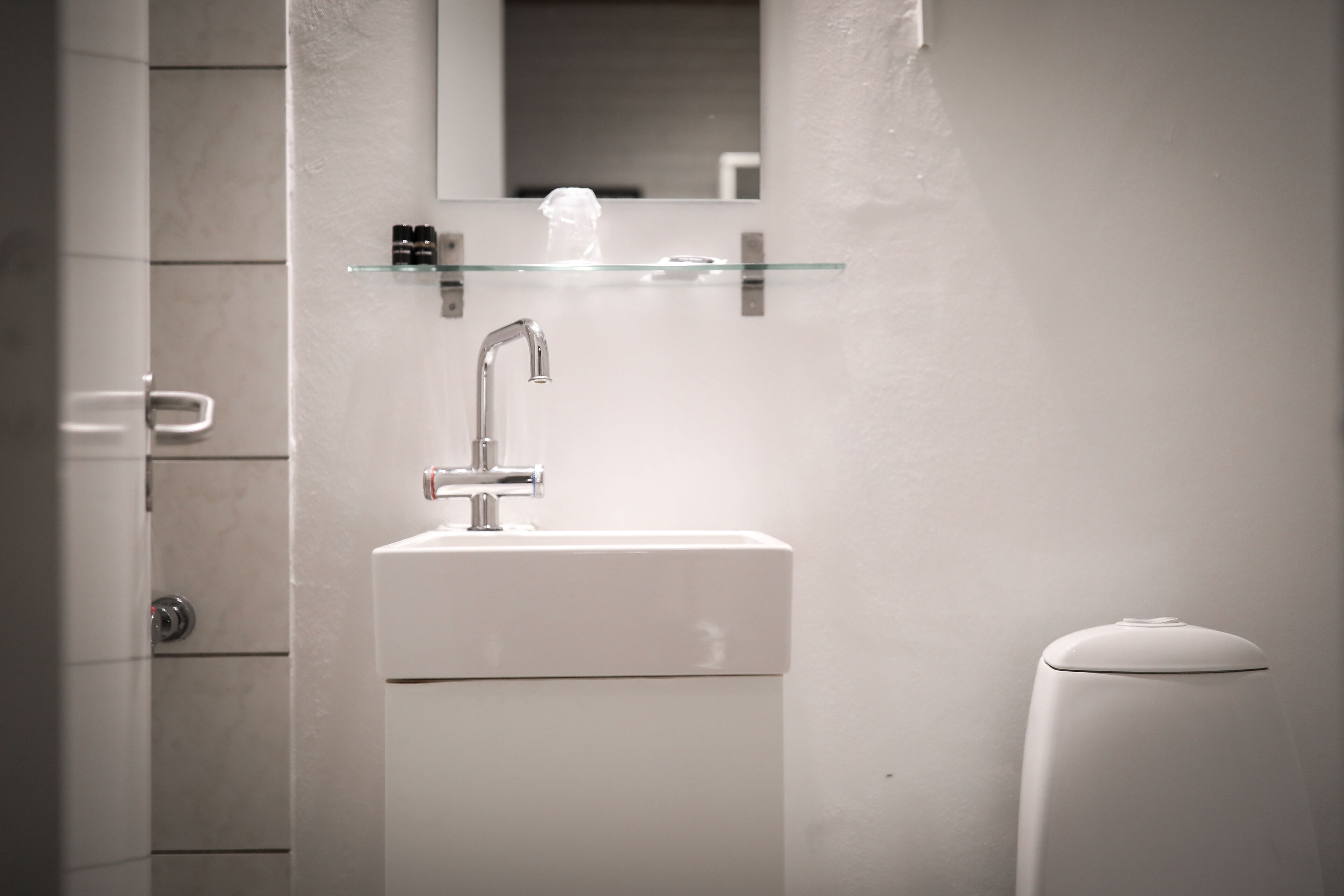 Un baño con un lavamanos blanco, un espejo encima y un estante de vidrio con artículos de aseo.