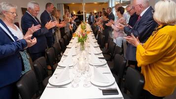 A formal dining setting with a long table set for a meal, surrounded by attendees clapping.