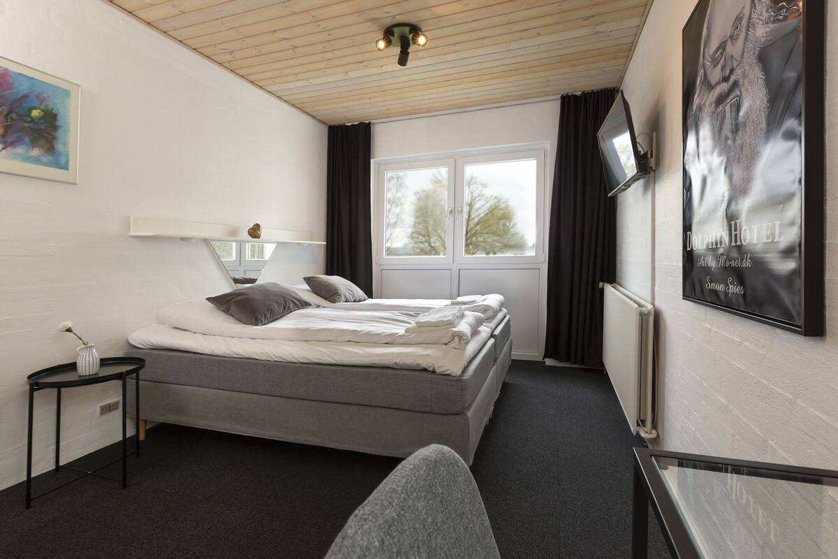 A hotel room with a bed, a small table with a vase, a window with curtains, and a poster on the wall.
