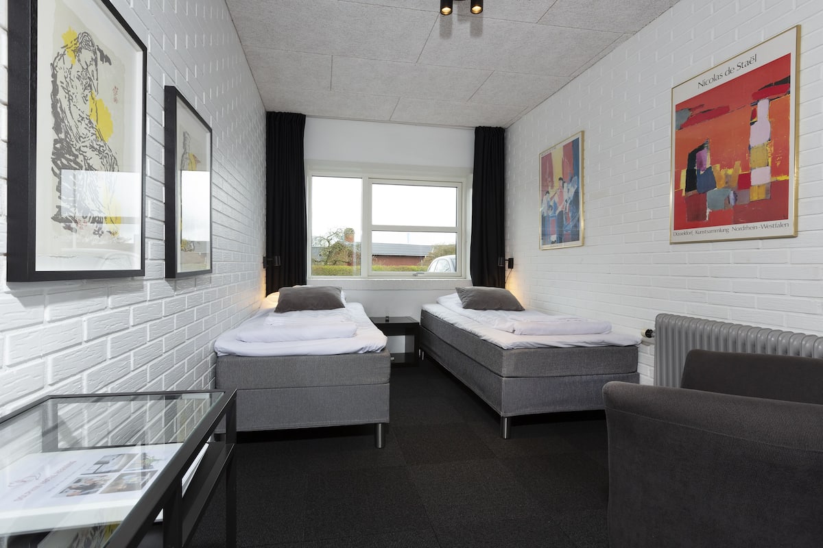 A hotel room with two single beds, a glass table, a chair, and artwork on the walls.
