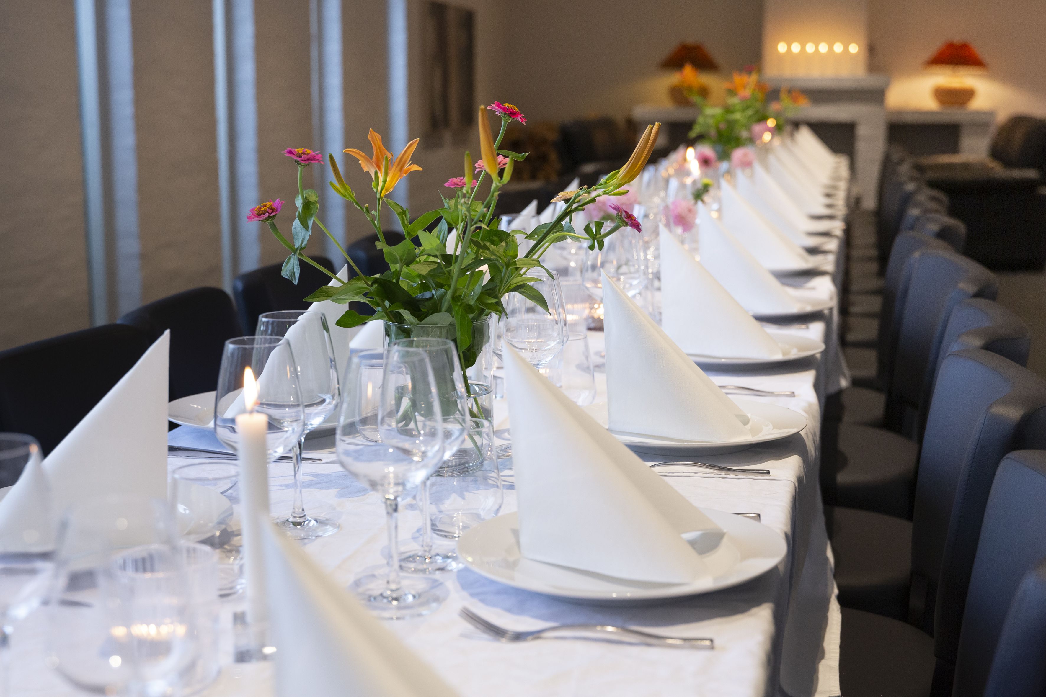 A long dining table set with white tablecloths, napkins, and glassware, with floral centerpieces and candles.