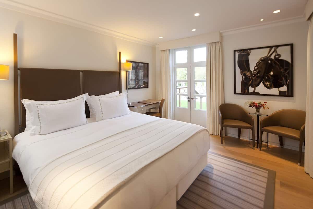 A hotel room with a large bed, a desk, two chairs, a lamp, and a window with curtains.