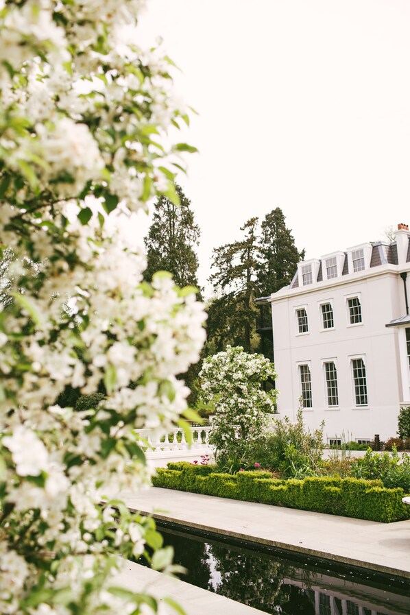 A white building with multiple windows and a garden with a water feature. - Coworth Park - Dorchester Collection (Ascot)