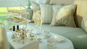 A tea service set on a table with a tiered tray, teapots, cups, and saucers, and a sofa with patterned cushions in a bright room with a view. - Coworth Park - Dorchester Collection (Ascot)