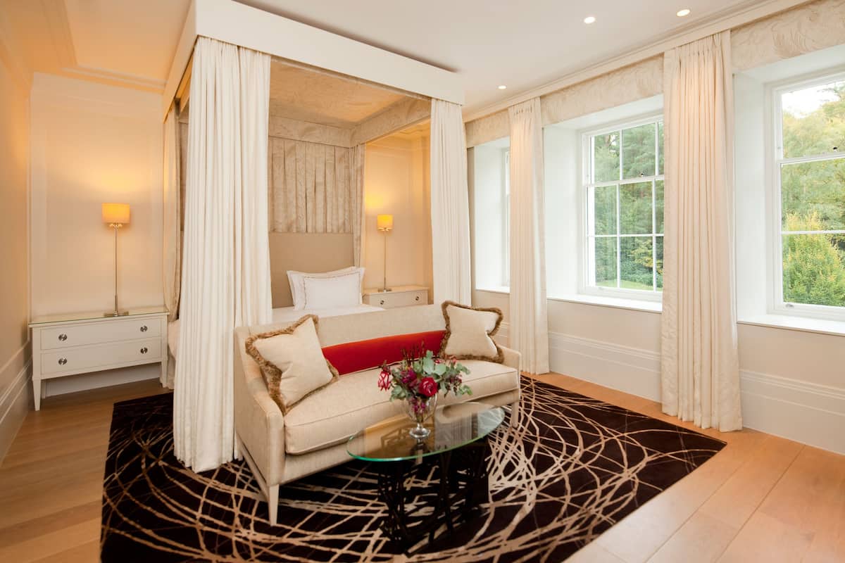 A bedroom with a four-poster bed, a sofa, a glass coffee table, and a view of greenery outside.