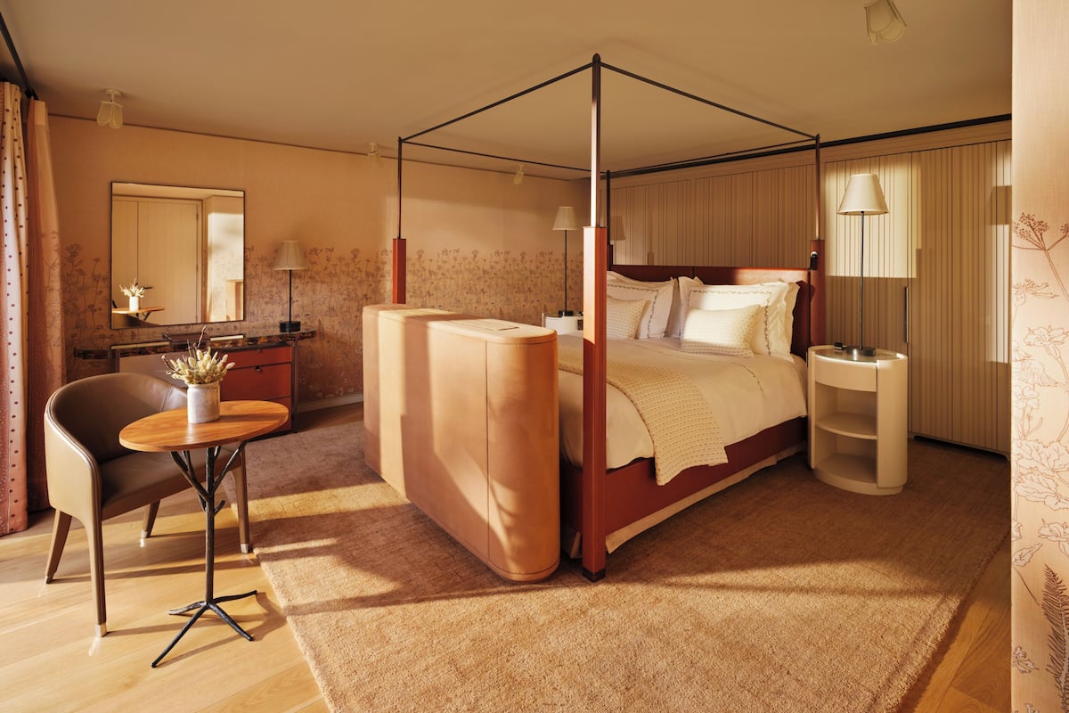 A four-poster bed with a canopy, a sitting area with a chair and table, a bedside lamp, and a mirror on the wall.