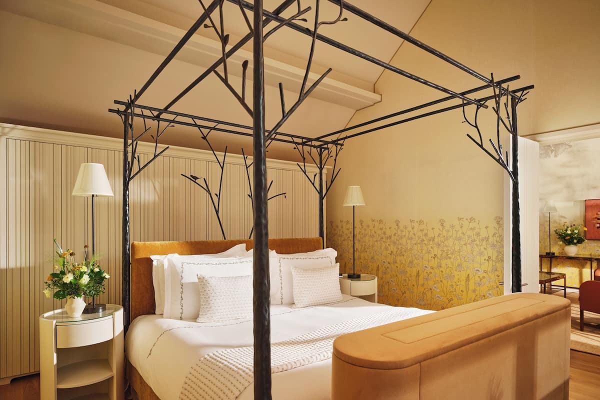 A four-poster canopy bed with white linens, two bedside tables with lamps, a wooden headboard, and a floral wallpaper pattern.