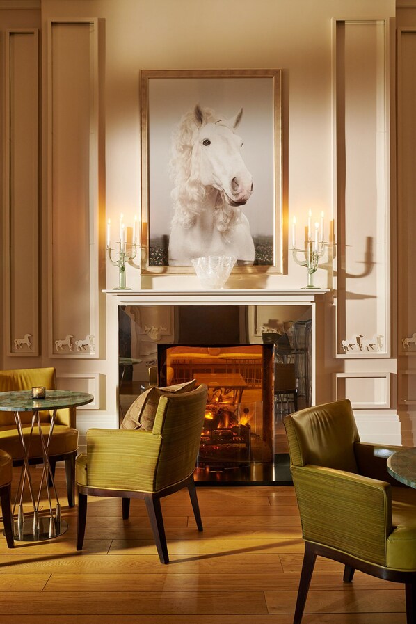 A room with a fireplace, two chairs, a table, and a framed horse portrait above the mantel. - Coworth Park - Dorchester Collection (Ascot)