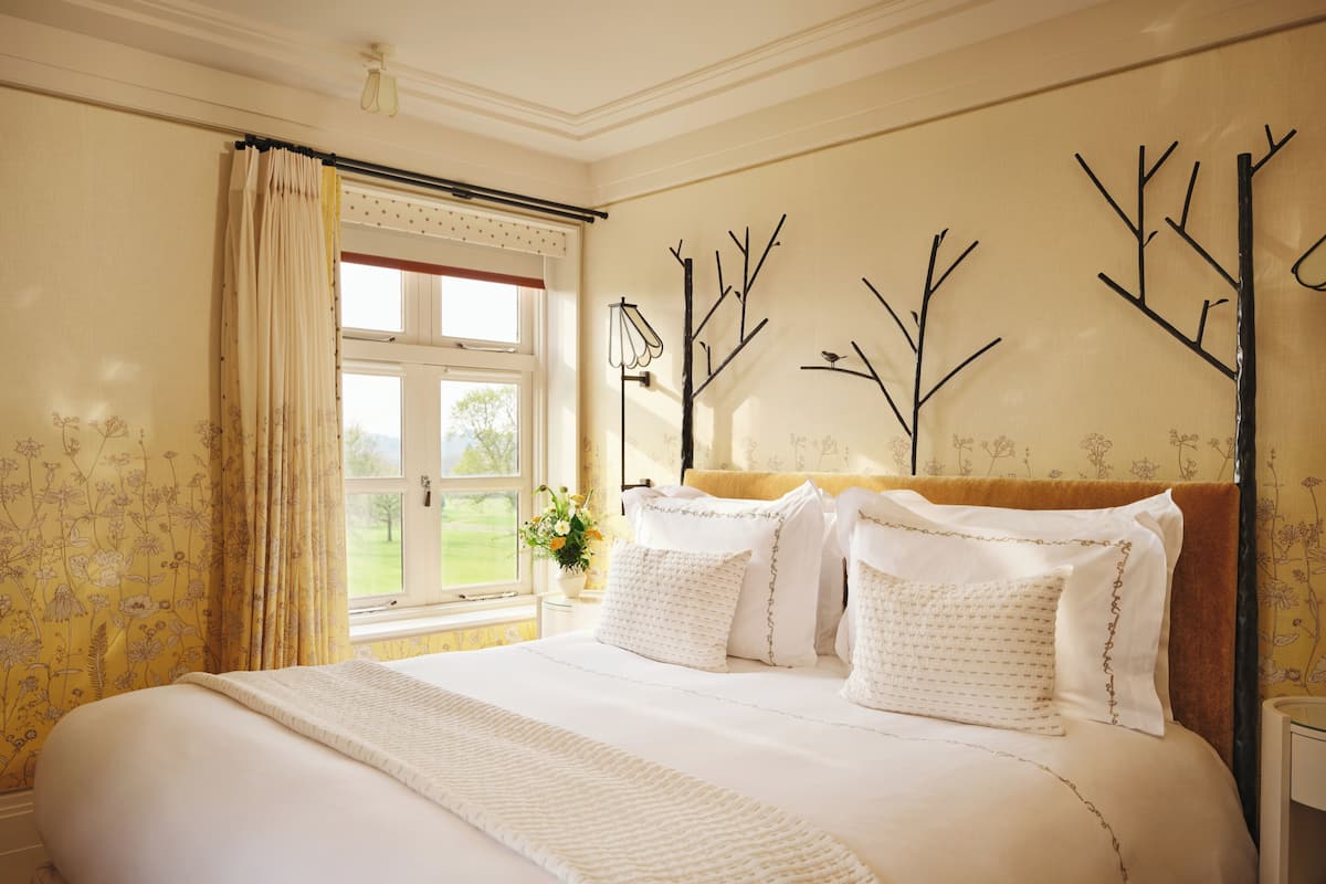 A neatly made bed with white linens, a window with curtains, and a wall with a branch decoration.