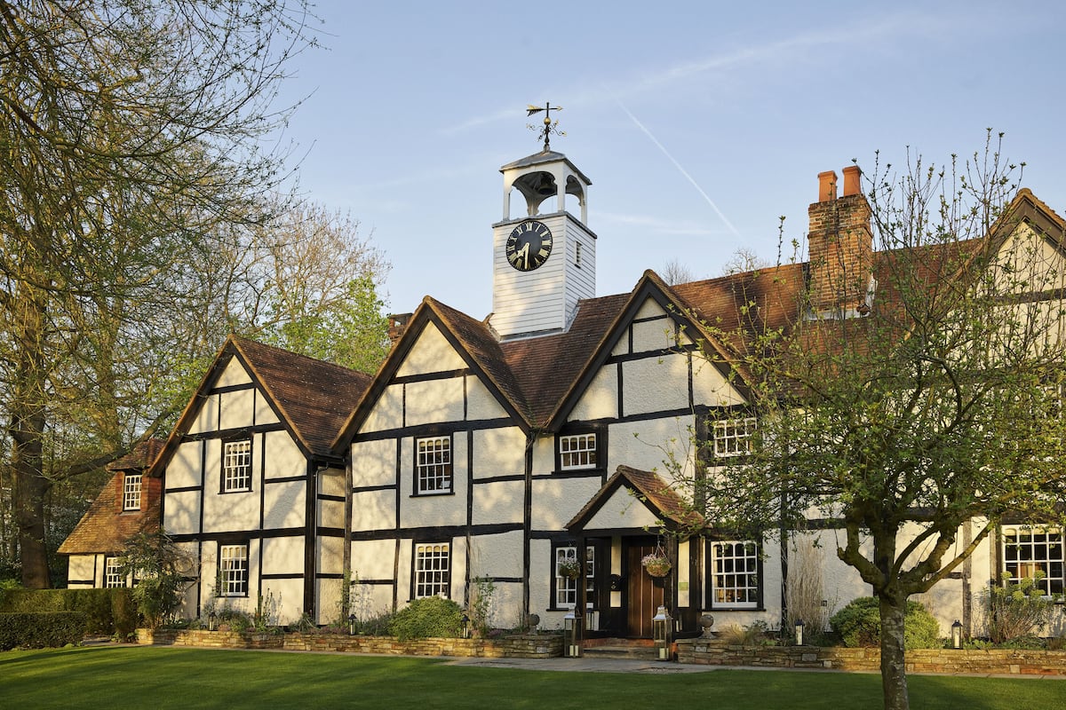 A traditional Tudor-style building with a prominent clock tower, a well-maintained lawn, and stone walls.