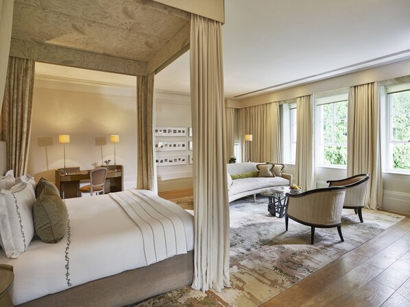 A spacious bedroom with a large bed, a sitting area with a sofa and chairs, a desk, and a view of greenery through the windows. - Coworth Park - Dorchester Collection (Ascot)