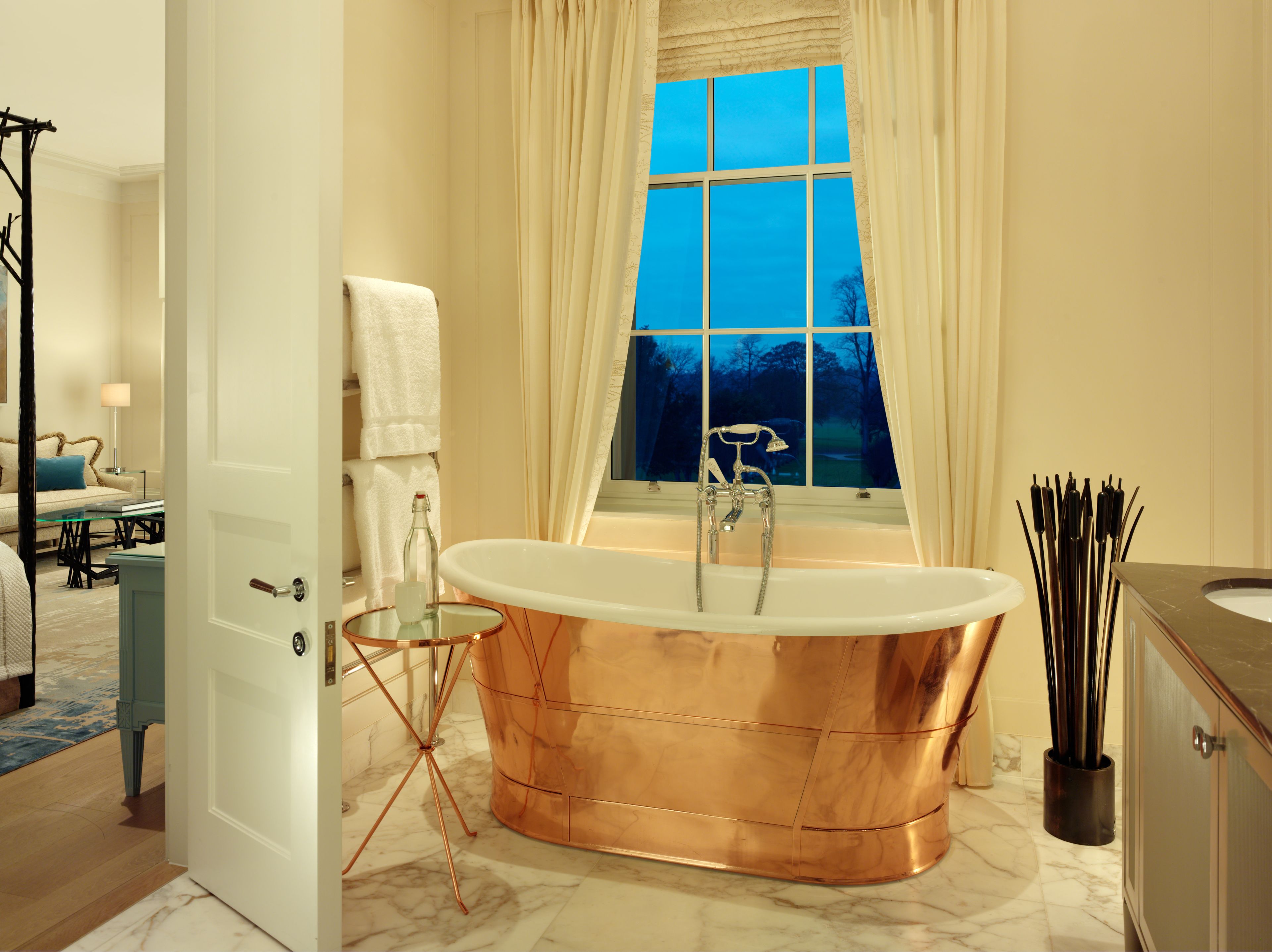 A modern bathroom with a freestanding copper bathtub, a glass-topped vanity, and a view of the outdoors through a large window.