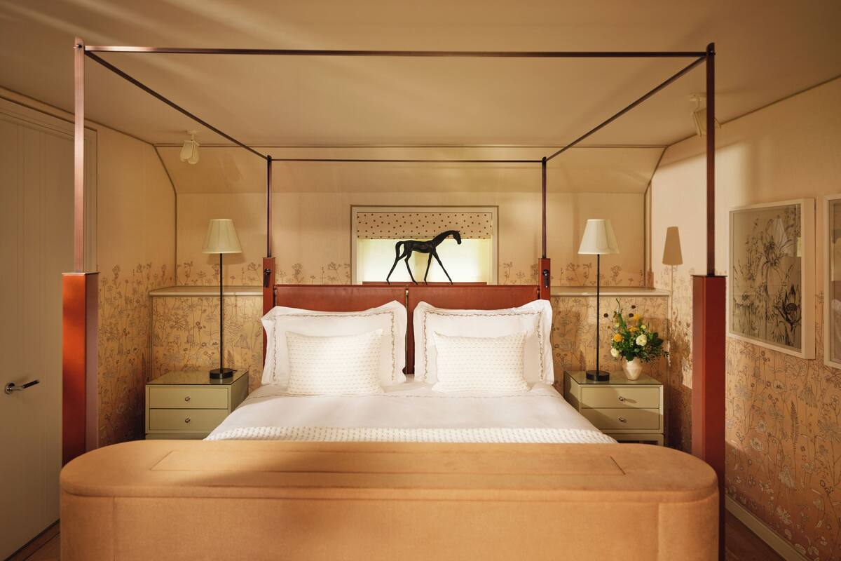 A bedroom with a canopy bed, two bedside tables, a nightstand, a wall-mounted lamp, and a floral wallpaper pattern.