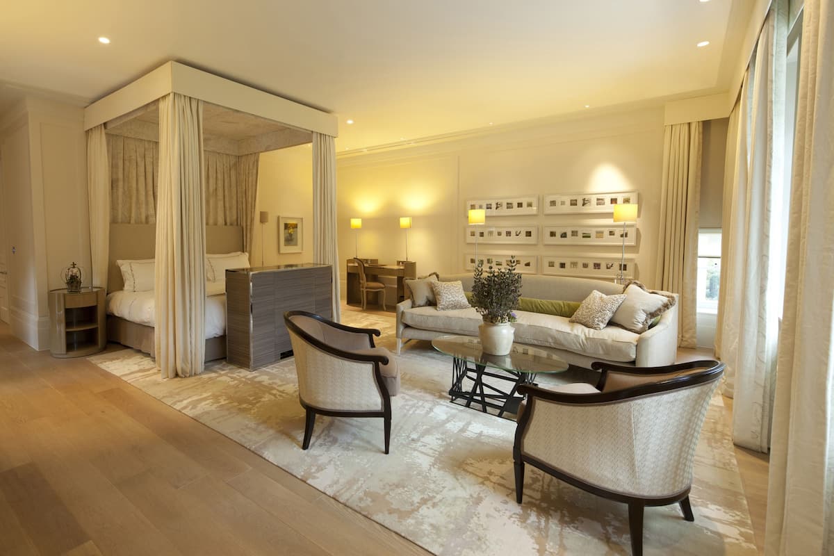 A modern hotel room with a large bed, a sitting area with a sofa and chairs, a dining table, and a large window with curtains.