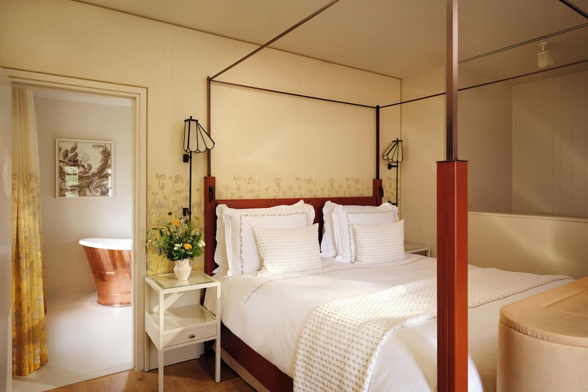 A four-poster bed with white linens, a bedside table with a vase of flowers, and a wall-mounted lamp.