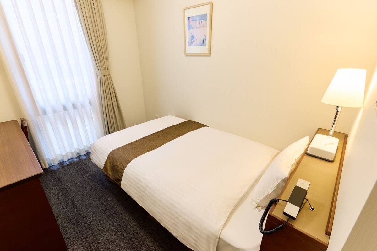 A hotel room with a single bed, a bedside table with a lamp, a telephone, and a window with curtains.