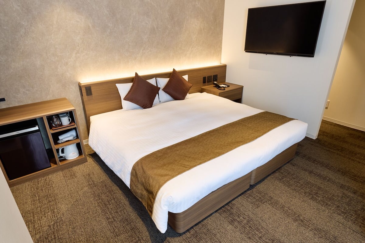 A hotel room with a large bed, a bedside table with a phone, a TV mounted on the wall, and a built-in storage unit with a dishwasher.
