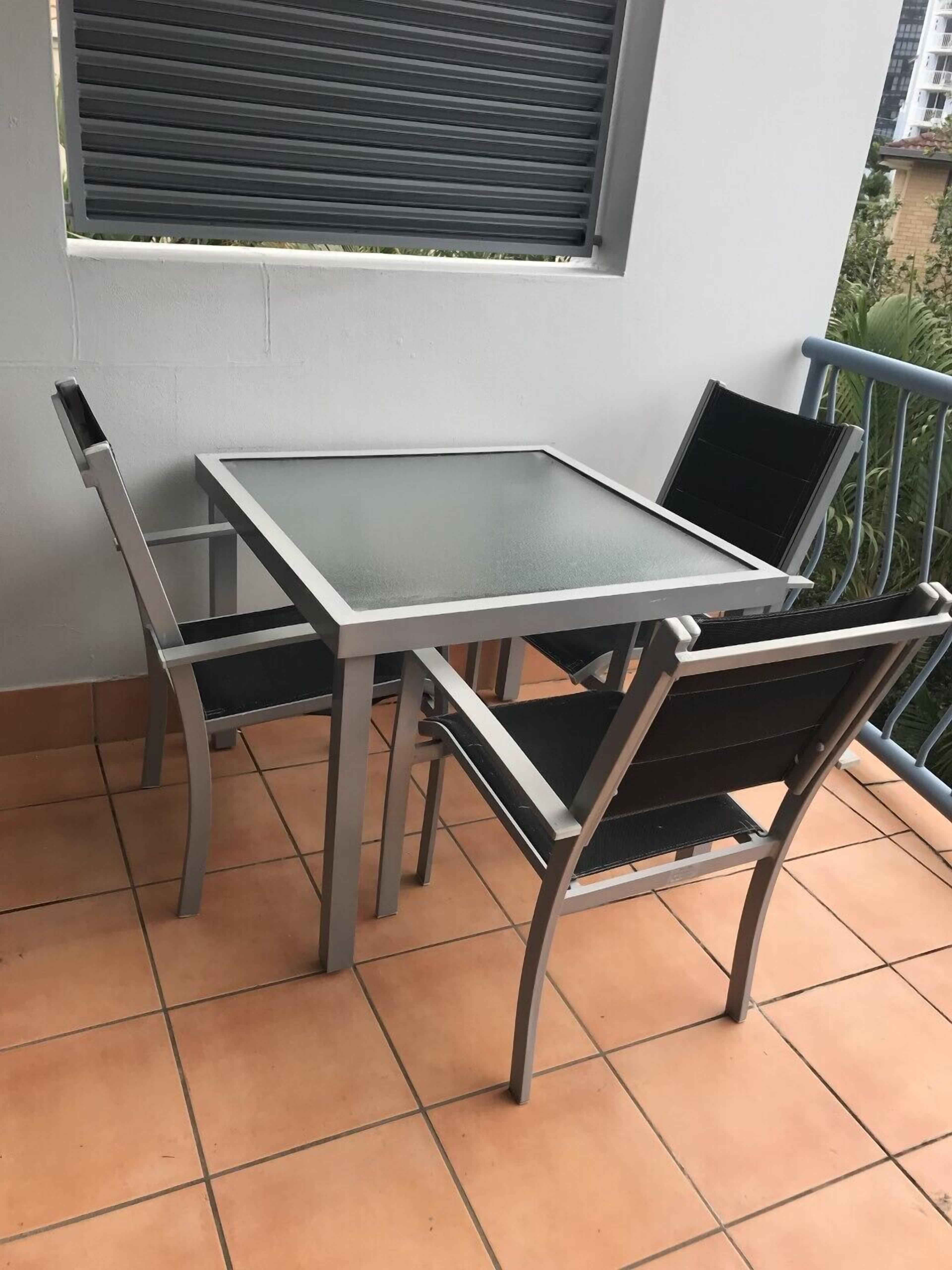 Uma varanda com mesa e cadeiras, uma janela com persianas e vista para um edifício.