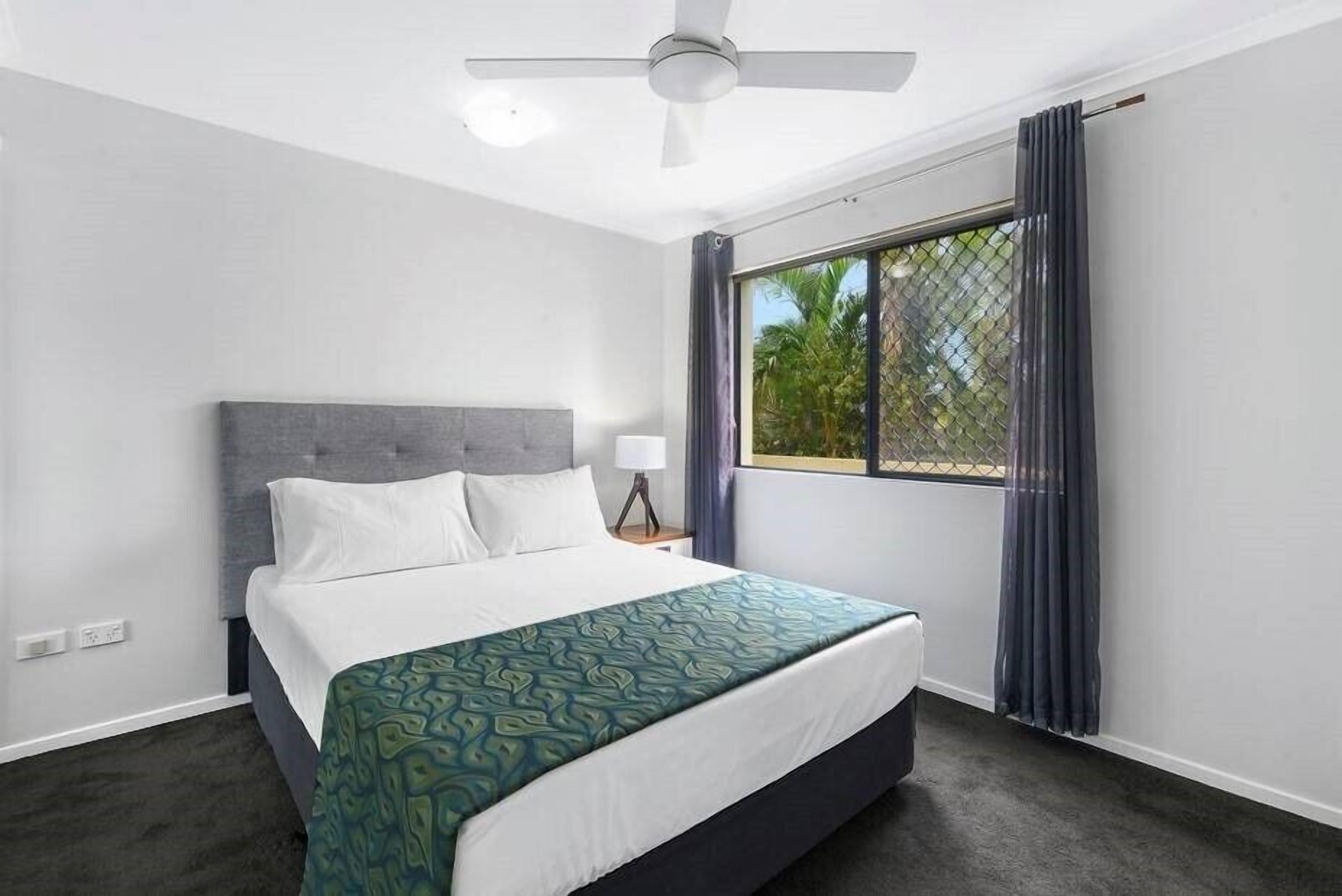 A hotel room with a bed, a ceiling fan, a window with curtains, and a view of greenery outside.