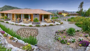 A house with a tiled roof, a covered patio with wicker furniture, and a garden with various plants and flowers. - Killawasi Lodge (Yanque)