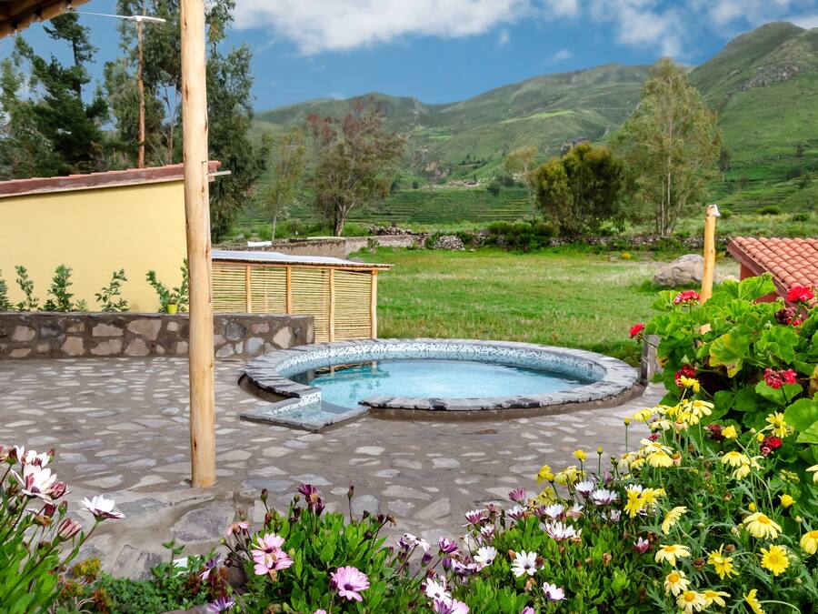 A hot tub surrounded by a stone patio, with a yellow building and a view of green hills in the background.