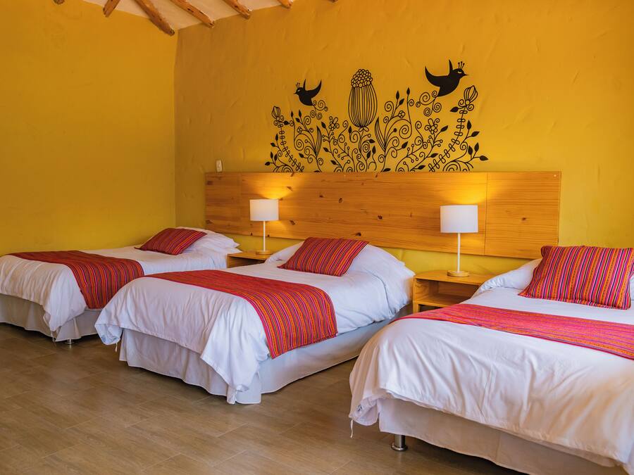 A hotel room with three beds, each with white bedding and red striped blankets. There are wooden headboards and bedside tables with lamps.