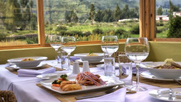 A dining table set with plates of food, glasses of water, and a scenic mountain view through large windows. - Killawasi Lodge (Yanque)