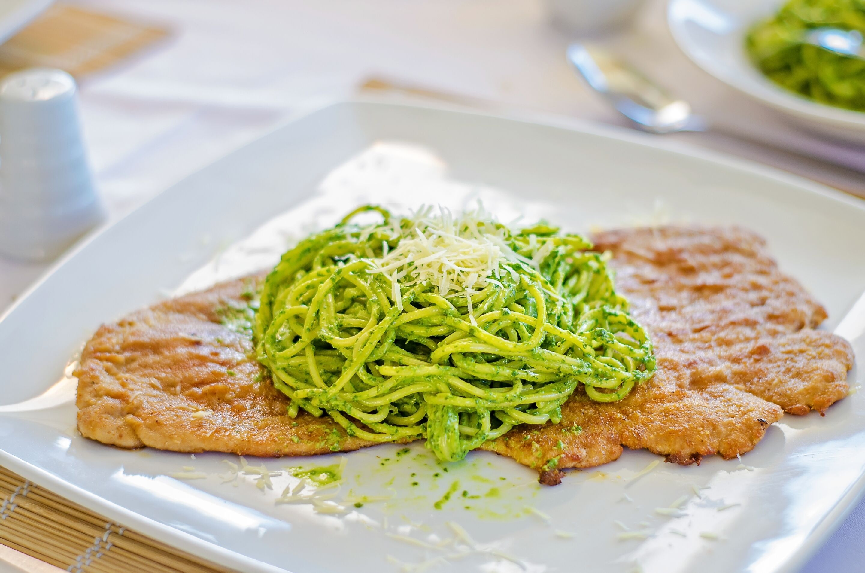 A plate of spaghetti with a green sauce and grated cheese, accompanied by two pieces of breaded chicken.