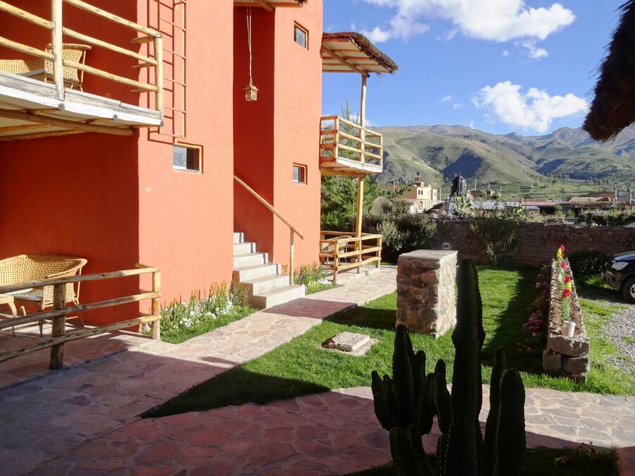 A red building with a staircase leading to a wooden deck, surrounded by a garden with cacti and flowers, and a view of distant mountains.
