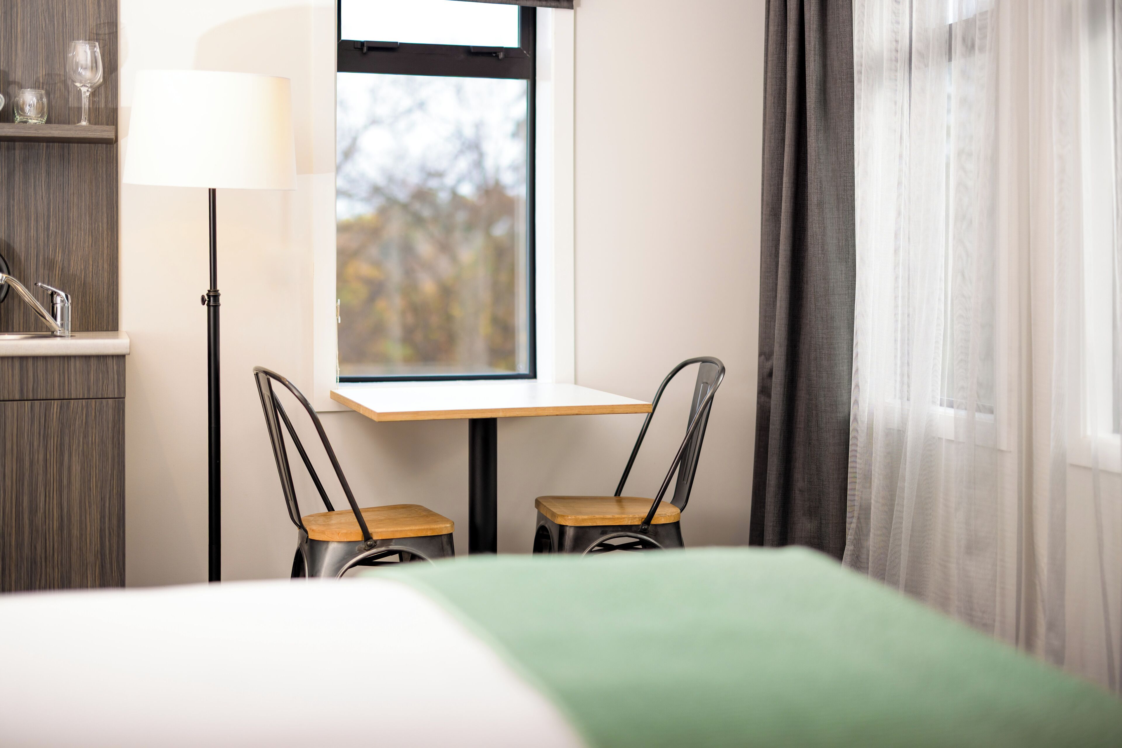 A hotel room with a bed, a desk with two chairs, a lamp, and a window with curtains.