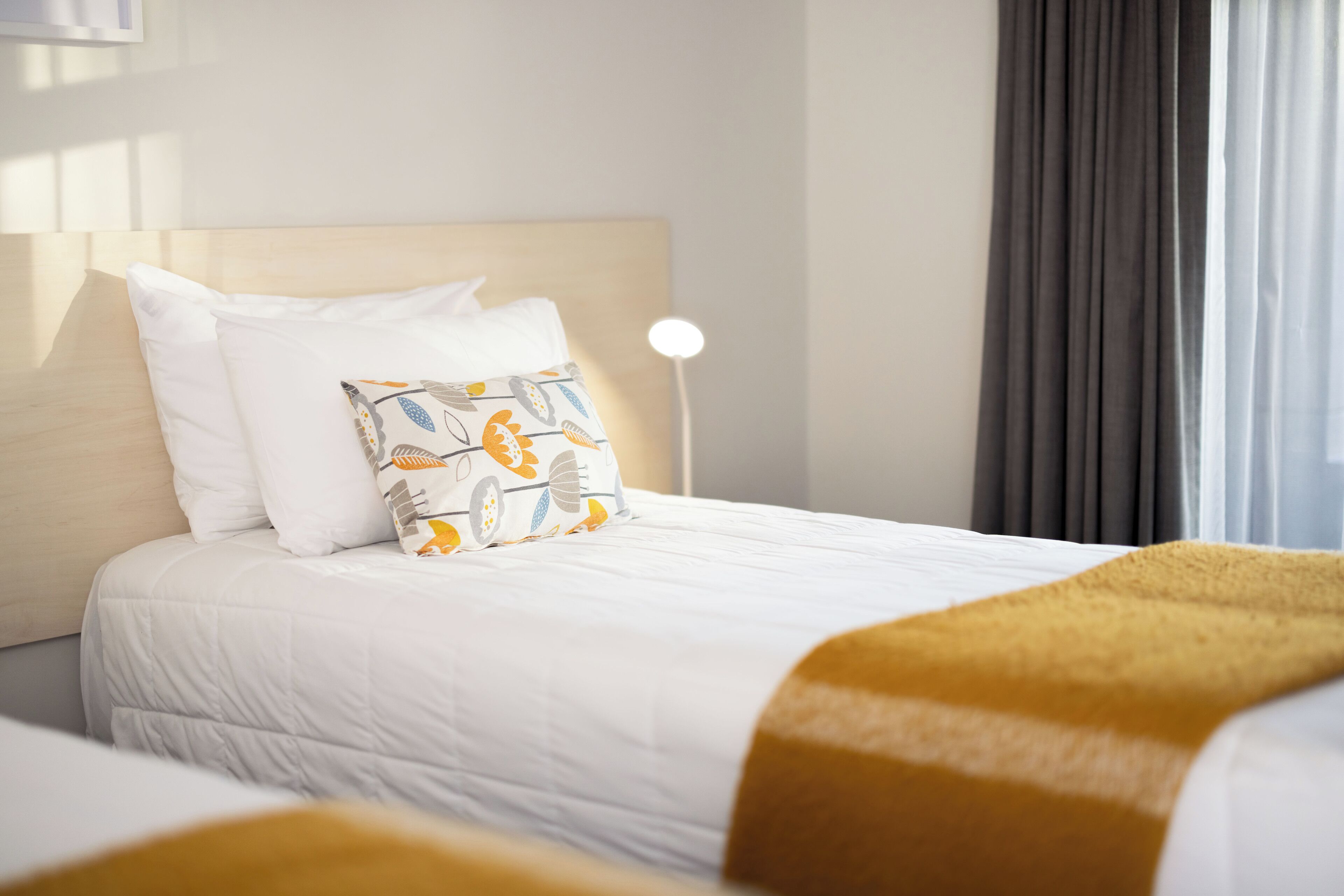A neatly made bed with white bedding and a patterned pillow, a bedside lamp, and a window with curtains.