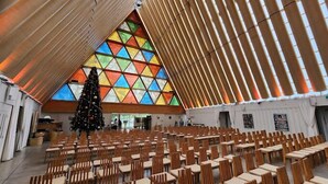 A large hall with a triangular stained glass window, wooden chairs, and tables arranged for an event. - Hotel Give (Christchurch)