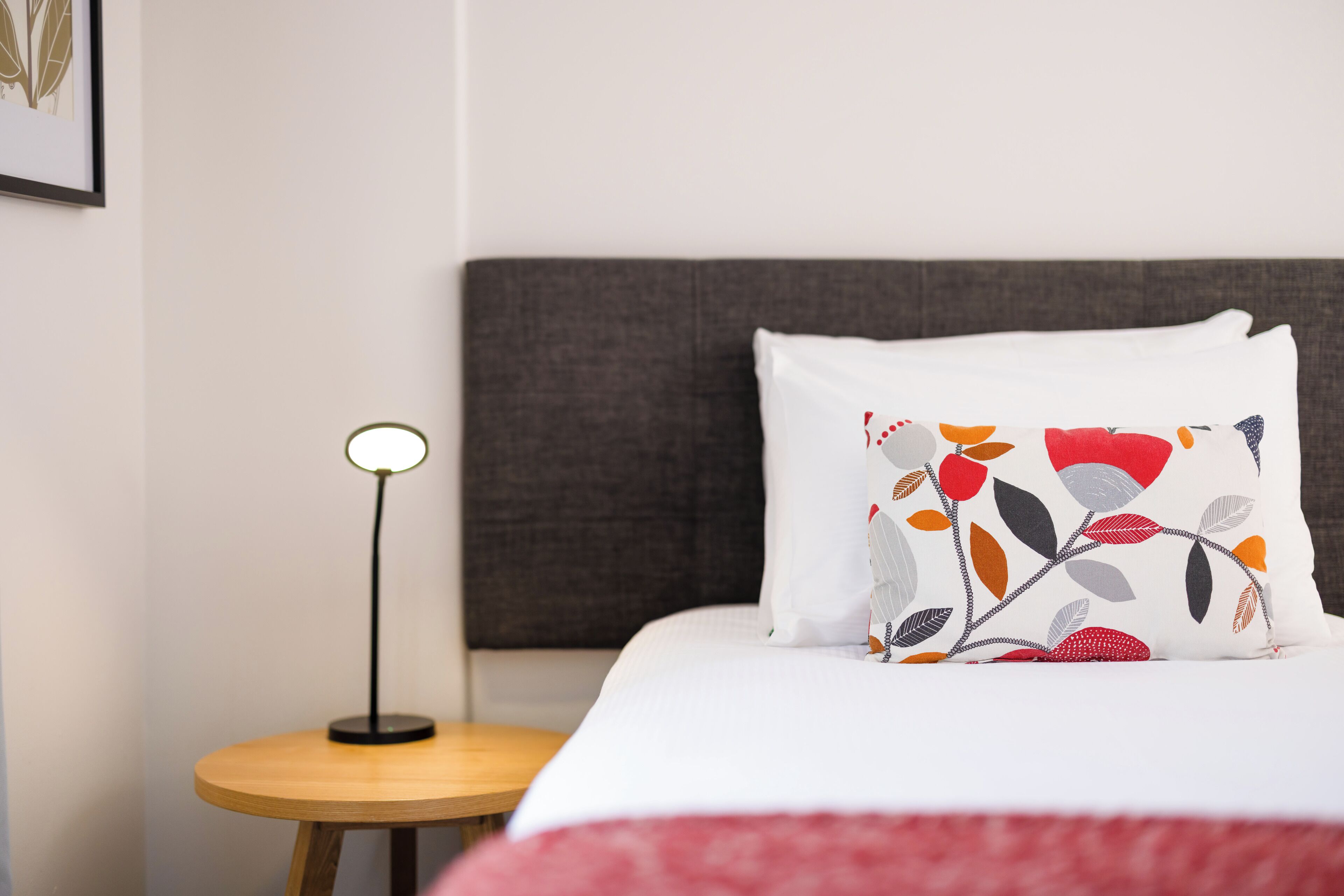 Una cama bien hecha con un almohadón blanco estampado con un diseño floral, una mesita de noche redonda de madera con una lámpara y un cabecero oscuro.