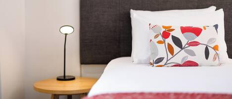 Una cama bien hecha con un almohadón blanco estampado con un diseño floral, una mesita de noche redonda de madera con una lámpara y un cabecero oscuro.