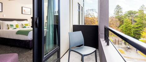 Habitación de hotel con cama, una silla y balcón con vistas a árboles y a una calle.