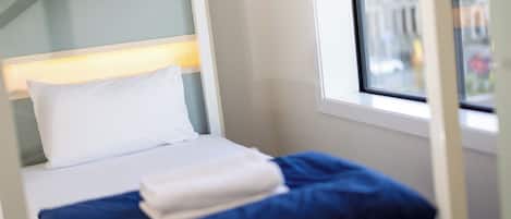 A single bed with a white pillow and blue bedding, a window with a view of buildings, and a white headboard.