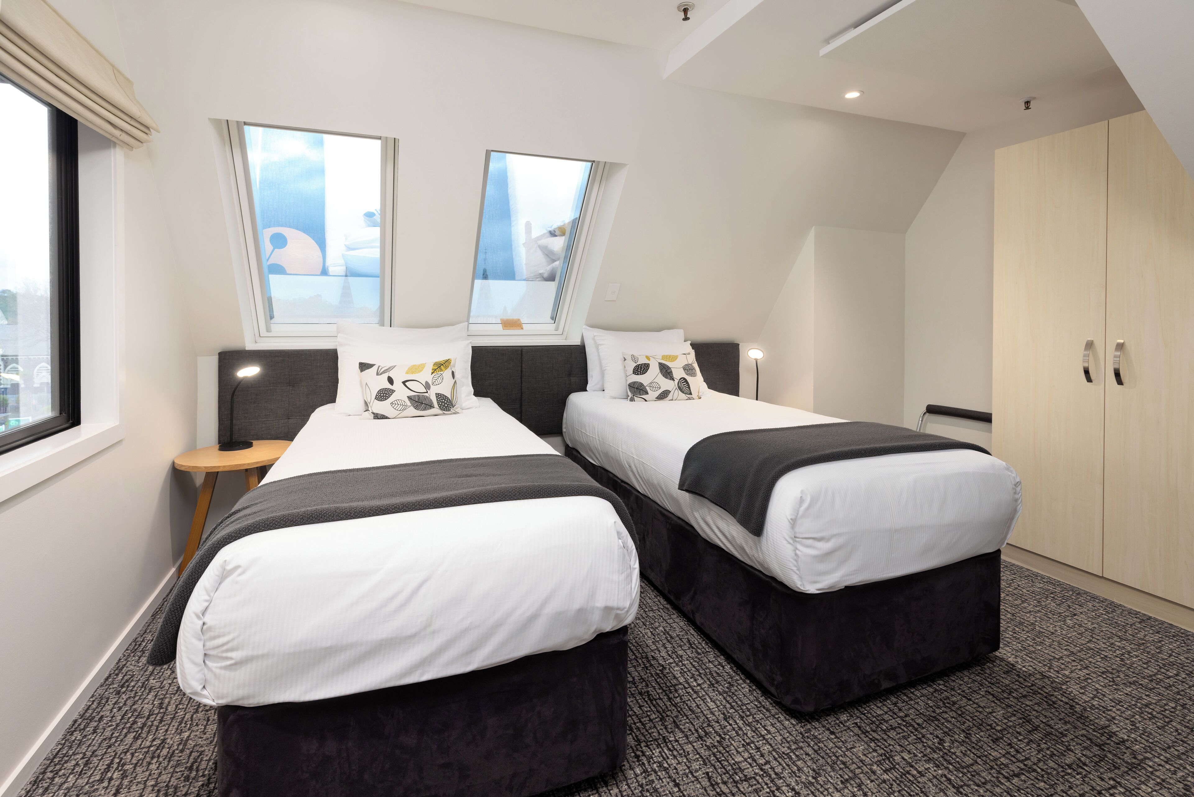 Una habitación de hotel moderna con dos camas, un tragaluz, un armario y una mesita auxiliar.