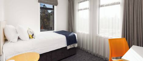 A hotel room with a bed, a small table, an orange chair, and large windows with curtains.