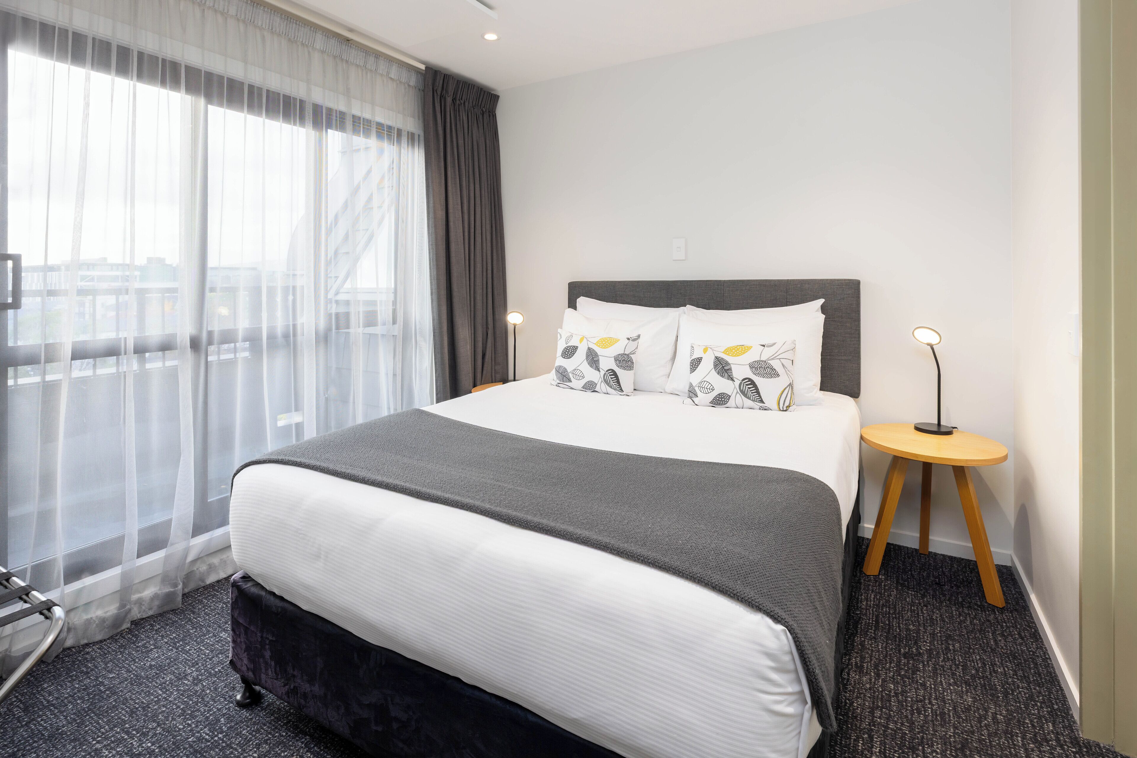 Quarto de hotel com uma cama grande, uma mesinha de cabeceira pequena, uma cadeira e vista da cidade através de cortinas leves.